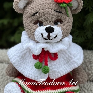 May include: A crocheted teddy bear wearing a white cape with a red ribbon and green accents, a red dress with green and white trim, and a red flower on its head.