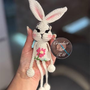 May include: White crocheted bunny with pink ears and a pink flower embroidered on its chest. The bunny has long legs with white balls at the ends. The bunny is being held by a hand.