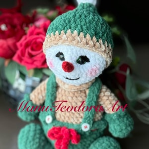 May include: A crocheted snowman toy with a green hat, white face, red nose, and red bow. The snowman is wearing a brown and green outfit with white buttons. The toy is sitting on a table with a blurred background.