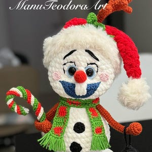 May include: A white crocheted snowman wearing a red and white Santa hat, a green and red scarf, and holding a candy cane and a small lantern. The snowman has a blue mouth, red nose, and black eyes.