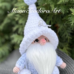 Peut inclure: Un gnome en crochet avec un chapeau et une barbe blancs, portant une tenue bleu clair et tenant une boule blanche. Le gnome est assis sur une surface en paille tressée. Le texte "Manu Teodora Art" est visible en arrière-plan.