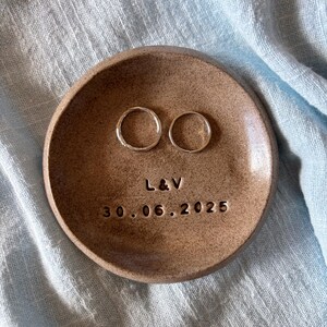 以下が含まれることがあります： 2つの銀色の指輪が置かれた、小さくて丸い茶色のセラミックリング皿。「L & V」のイニシャルと日付「30.06.2025」が刻まれています。皿は水色の布の上に置かれています。