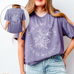 May include: Heather purple t-shirt with the text "THIS IS MY STORY, THIS IS MY SONG" in a circular design around a floral graphic. The back of the shirt has the text "PRAISING MY SAVIOR ALL THE DAY LONG" in a circular design around a floral graphic.