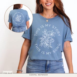 May include: Steel blue t-shirt with white floral design and text "THIS IS MY STORY, THIS IS MY SONG". The back of the shirt has the text "PRAISING MY SAVIOR ALL THE DAY LONG". The shirt is part of the Gospel Repwear collection.