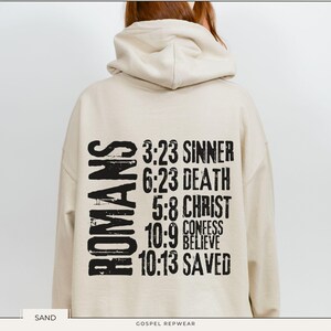 May include: A beige hoodie with a black graphic print on the back that reads "ROMANS 3:23 SINNER 6:23 DEATH 5:8 CHRIST 10:9 CONFESS BELIEVE 10:13 SAVED".