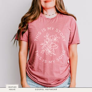 May include: Heather mauve t-shirt with the text "THIS IS MY STORY, THIS IS MY SONG" in a circular design around a floral illustration. The shirt is a short-sleeved crew neck.