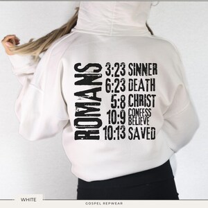 May include: White hoodie with black text that reads "ROMANS" and bible verses, including "3:23 SINNER", "6:23 DEATH", "5:8 CHRIST", "10:9 CONFESS BELIEVE", and "10:13 SAVED".