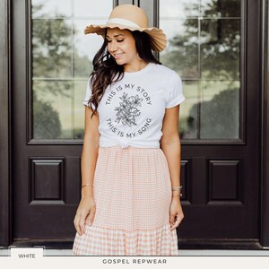 May include: White t-shirt with the text "THIS IS MY STORY, THIS IS MY SONG" encircling a floral design. The shirt is paired with a peach and white gingham skirt and a straw hat. The image is set against a dark brown door.