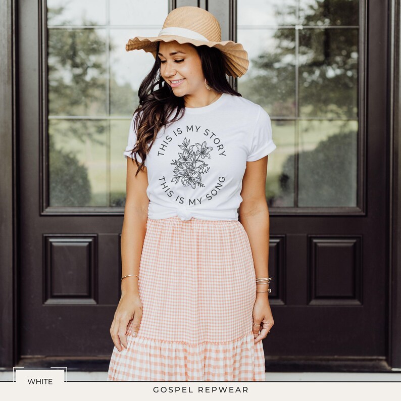 May include: White t-shirt with the text "THIS IS MY STORY, THIS IS MY SONG" encircling a floral design. The shirt is paired with a peach and white gingham skirt and a straw hat. The image is set against a dark brown door.