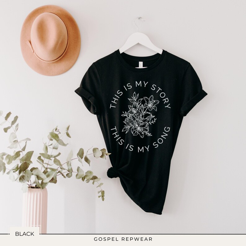 May include: Black t-shirt with the text "THIS IS MY STORY, THIS IS MY SONG" in a white circular design with floral accents. The shirt is hanging on a white hanger. A tan hat is visible in the background.