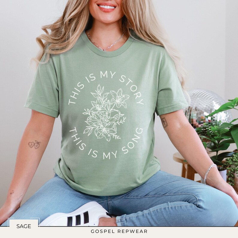 May include: Sage green t-shirt with the text "THIS IS MY STORY, THIS IS MY SONG" in a circular design around a floral illustration. The shirt is a crew neck and is paired with blue jeans.