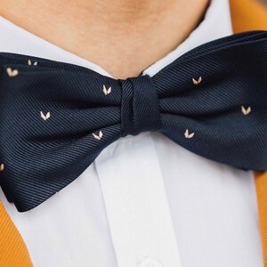 May include: A navy blue bow tie with small gold accents in the shape of a heart. The bow tie is tied and worn on a white shirt.