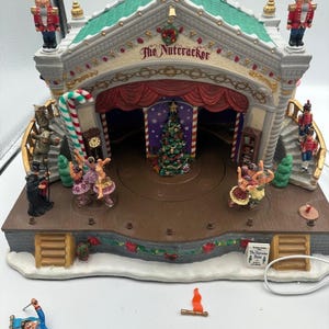 Peut inclure: Une scène miniature sur le thème de Noël, avec un bâtiment au toit vert portant l'inscription "The Nutcracker" au-dessus d'un rideau rouge. La scène comprend un sapin de Noël, des figurines et une canne en bonbon. Des figurines supplémentaires sont éparpillées autour de la base.