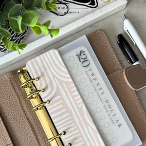 May include: A beige leather bound planner with gold rings and a clear plastic pocket containing a savings challenge tracker. The tracker is labelled "£20 Twenty Pound Savings Challenge I Save £1.200".