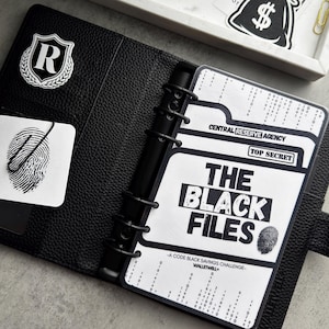 May include: A black leather planner, open to a page with the text "THE BLACK FILES." The planner has a black cover with a white "R" emblem and a fingerprint graphic. A tray with gold paper clips and business cards is in the background.