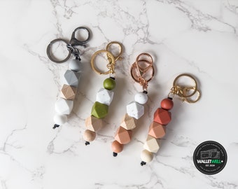 May include: Four colorful geometric bead keychains with gold and silver key rings. The beads are made of wood and silicone and come in shades of gray, green, brown, and white.