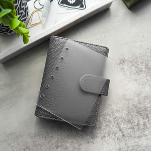 May include: A gray leather bound planner with a clear plastic cover and six silver rings. The planner has a flap closure with a silver button.