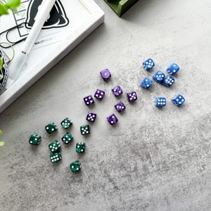 Può includere: Vista dall'alto di dadi verdi, viola e blu, ciascuno con puntini bianchi. Sono visibili anche una penna bianca e un'immagine incorniciata con la scritta "CASH KD WINNER", oltre a un organizer verde.