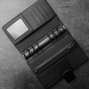 May include: Black leather binder, open to reveal a textured interior. It has a clear window, card slots, a zipped pocket, and a snap closure. The binder incorporates a metal ring mechanism for securing pages.