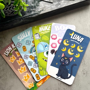 Può includere: Una collezione di carte sfida di risparmio colorate con personaggi dei cartoni animati e design a tema. Le carte includono illustrazioni di un leone, Sully, Mike, Boo e Luna, con importi come 5 €, 10 € e 20 €. Una piccola pianta e un vassoio sono sullo sfondo.