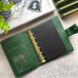 May include: A green leather bound planner with gold rings and a black insert. The planner is open and the rings are visible. The planner has a clear plastic pocket on the front cover.