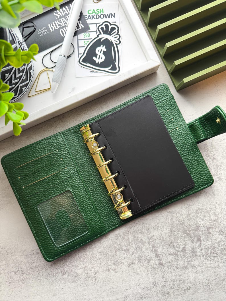 May include: A green leather bound planner with gold rings and a black insert. The planner is open and the rings are visible. The planner has a clear plastic pocket on the front cover.