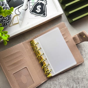 May include: An open beige leather bound planner with gold rings and a clear plastic pocket insert. The planner is open to a blank white page. The planner is on a gray surface with a green organizer and a sticker of a money bag with a dollar sign on it.