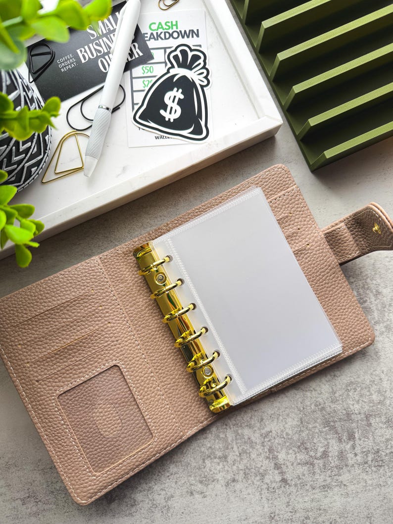 May include: An open beige leather bound planner with gold rings and a clear plastic pocket insert. The planner is open to a blank white page. The planner is on a gray surface with a green organizer and a sticker of a money bag with a dollar sign on it.