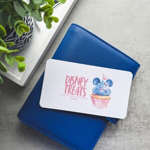 Op de afbeelding: Een blauwe leren portemonnee met een witte kaart met de tekst "Disney Treats" en een cupcake-illustratie. De kaart ligt bovenop de portemonnee, die op een grijze ondergrond ligt. Een dienblad met een plant staat op de achtergrond.