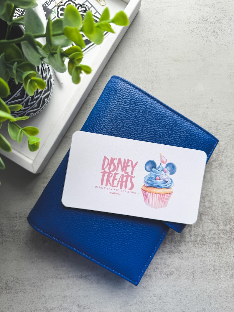 Op de afbeelding: Een blauwe leren portemonnee met een witte kaart met de tekst "Disney Treats" en een cupcake-illustratie. De kaart ligt bovenop de portemonnee, die op een grijze ondergrond ligt. Een dienblad met een plant staat op de achtergrond.