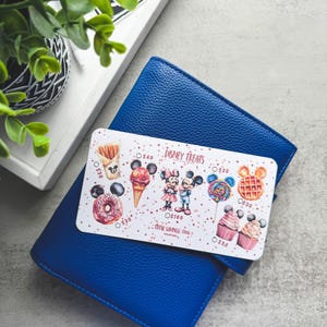 Op de afbeelding: Een blauwe leren portemonnee met een Disney Treats-kaart met illustraties van Mickey en Minnie Mouse met diverse lekkernijen. De kaart bevat tekst en prijskaartjes. Een plant en een dienblad zijn op de achtergrond.
