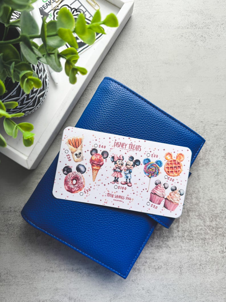 Op de afbeelding: Een blauwe leren portemonnee met een Disney Treats-kaart met illustraties van Mickey en Minnie Mouse met diverse lekkernijen. De kaart bevat tekst en prijskaartjes. Een plant en een dienblad zijn op de achtergrond.