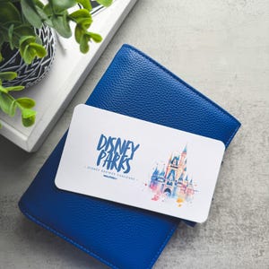 Op de afbeelding: Een blauwe leren portemonnee met een witte kaart erop. De kaart toont de tekst "DISNEY PARKS" en een aquarel illustratie van een kasteel. De portemonnee ligt op een grijze ondergrond, met een dienblad en een plant op de achtergrond.