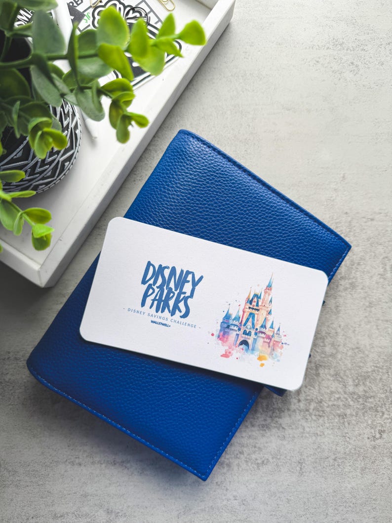 Op de afbeelding: Een blauwe leren portemonnee met een witte kaart erop. De kaart toont de tekst "DISNEY PARKS" en een aquarel illustratie van een kasteel. De portemonnee ligt op een grijze ondergrond, met een dienblad en een plant op de achtergrond.