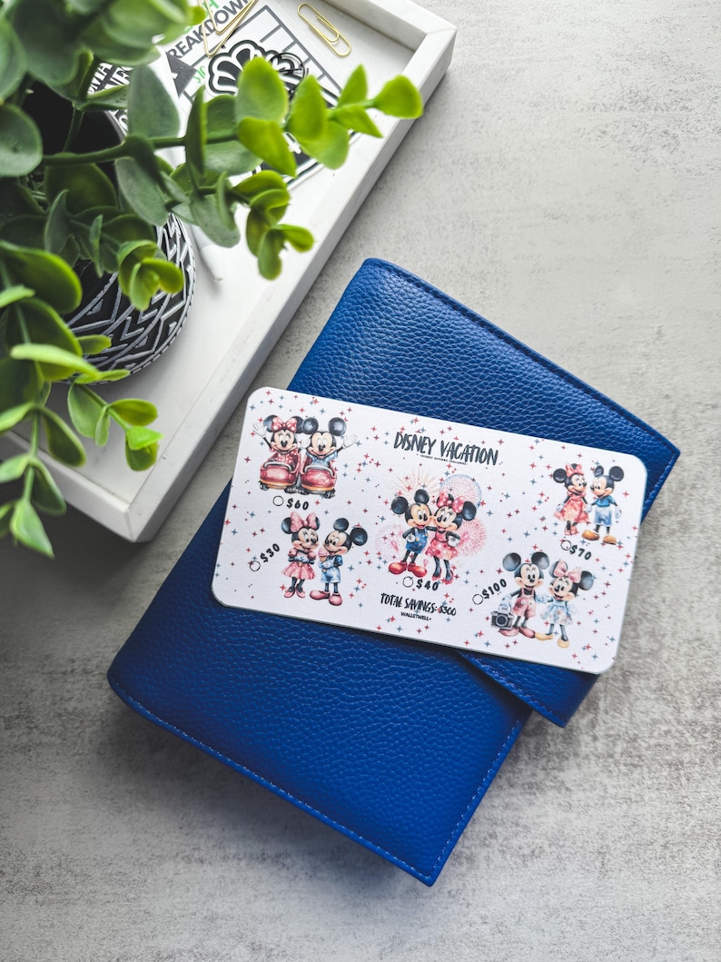 Op de afbeelding: Een blauwe leren portemonnee met een Disney Vacation spaartrackerkaart. De kaart bevat illustraties van Mickey en Minnie Mouse in verschillende outfits. De tekst "DISNEY VACATION" en spaarbedragen zijn zichtbaar.