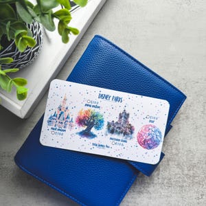 Op de afbeelding: Een blauwe leren portemonnee met een Disney Parks cadeaukaart erop. De cadeaukaart bevat illustraties van Disney park iconen, waaronder een kasteel en een kleurrijke boom. De kaart heeft de tekst "Disney Parks".