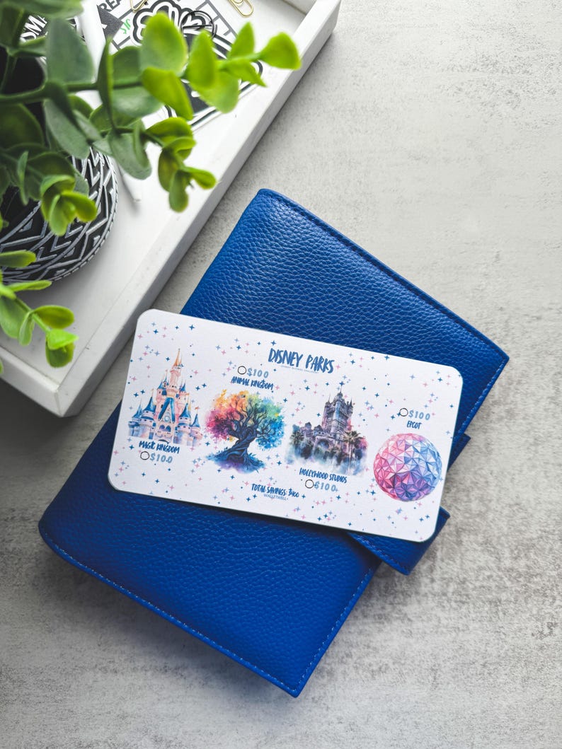 Op de afbeelding: Een blauwe leren portemonnee met een Disney Parks cadeaukaart erop. De cadeaukaart bevat illustraties van Disney park iconen, waaronder een kasteel en een kleurrijke boom. De kaart heeft de tekst "Disney Parks".