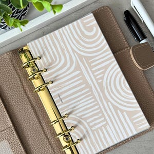 Puede incluir: Una agenda de cuero beige con anillas doradas y un inserto de papel con un patrón abstracto blanco y beige. La agenda tiene el texto "WALLETWELL+" impreso en la parte inferior.