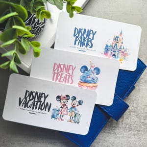 Op de afbeelding: Drie witte kaarten met Disney-ontwerpen liggen op een blauwe portemonnee. De kaarten tonen de zinnen "Disney Parks", "Disney Treats" en "Disney Vacation". Elke kaart heeft een illustratie, waaronder een kasteel, een cupcake en Mickey en Minnie Mouse met bagage.