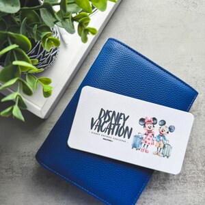 Op de afbeelding: Een blauwe leren portemonnee met een witte kaart met de tekst "Disney Vacation" en illustraties van Mickey en Minnie Mouse met koffers. Een kleine potplant en een dienblad staan op de achtergrond.