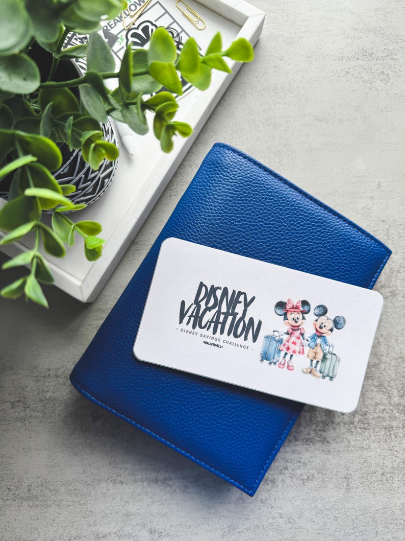 Op de afbeelding: Een blauwe leren portemonnee met een witte kaart met de tekst "Disney Vacation" en illustraties van Mickey en Minnie Mouse met koffers. Een kleine potplant en een dienblad staan op de achtergrond.