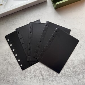 May include: Five black plastic page protectors with six holes down the side and a stitched edge. The protectors are stacked on top of each other.