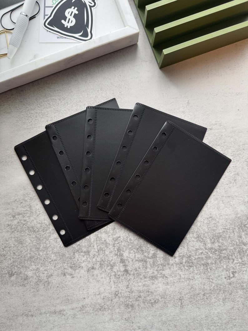 May include: Five black plastic page protectors with six holes down the side and a stitched edge. The protectors are stacked on top of each other.