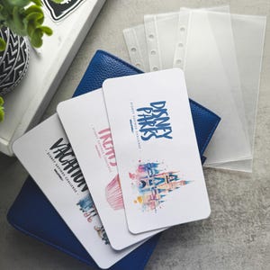 Op de afbeelding: Een blauwe leren portemonnee met drie witte kaarten met Disney-thema afbeeldingen en tekst. De kaarten lezen "Vacation", "Incity" en "Disney Parks Savings Challenge". Transparante plastic hoezen zijn zichtbaar op de achtergrond, samen met een decoratief dienblad.