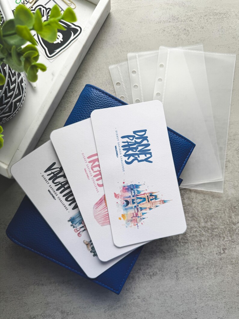 Op de afbeelding: Een blauwe leren portemonnee met drie witte kaarten met Disney-thema afbeeldingen en tekst. De kaarten lezen "Vacation", "Incity" en "Disney Parks Savings Challenge". Transparante plastic hoezen zijn zichtbaar op de achtergrond, samen met een decoratief dienblad.
