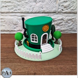 May include: A green leprechaun hat with a white picket fence and a small house on top. The hat is sitting on a green base.