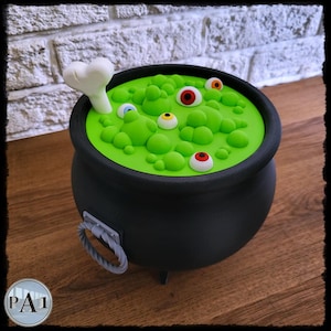 May include: A black cauldron filled with green liquid and floating eyeballs. A white bone sticks out of the green liquid. The cauldron has a handle and three small feet. The cauldron is on a wooden surface.