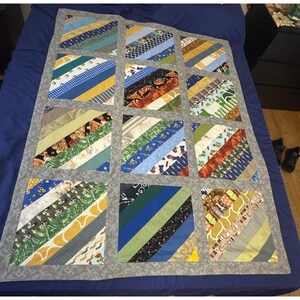 May include: A patchwork quilt with a grey border, featuring a grid of squares. Each square is composed of diagonal strips of various patterned fabrics in colours like blue, yellow, green, and brown. The quilt is laid out on a blue surface.