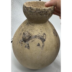 May include: A beige, antique pottery vessel with a rounded base and a smaller, cup-like top. The vessel has a dark, stylized animal figure painted on the front. The pottery has a textured, aged appearance.
