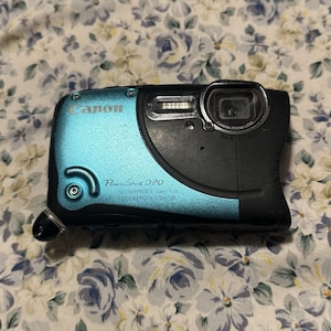 May include: A Canon PowerShot D20 digital camera. The camera is black and teal, with the Canon logo on the top left. The camera is waterproof up to 10m/33ft. The lens is labeled Canon Zoom Lens 5x IS.